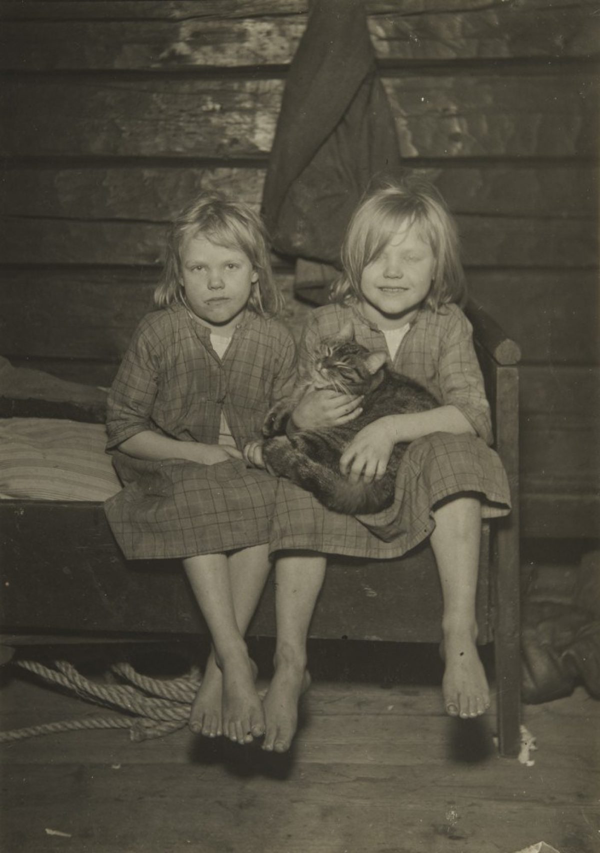 Tvillingflickorna Tyyne och Aili samt katten Matti på sängkanten i ett rökpörte i Pielavesi i Jokilahti på 1920-talet. Bild: Ahti Rytkönen/Museiverkets Bildsamlingar
