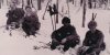 Mustavalkokuvassa neljä sotilasta hiihtovarusteineen istuu lumessa. Taustalla harvaa metsää.